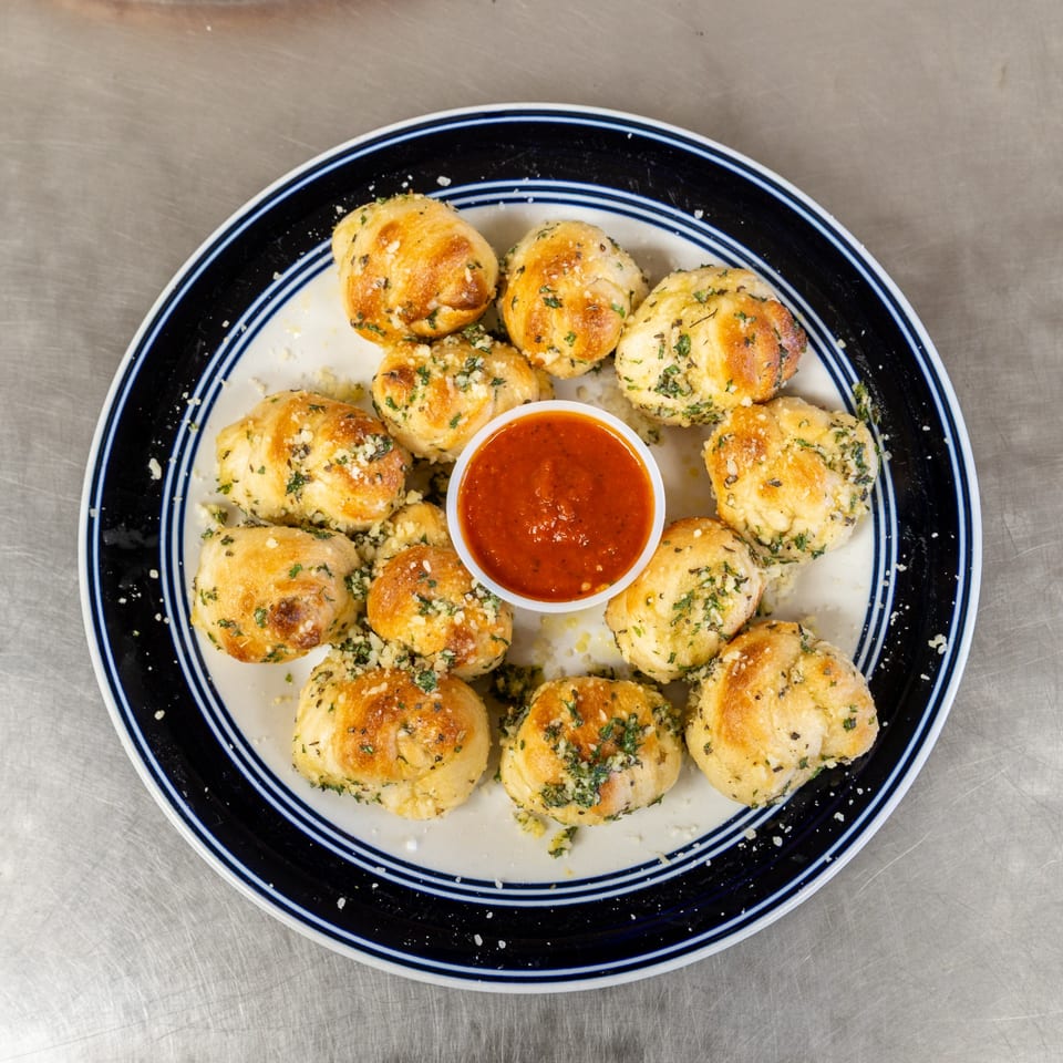 A plate of Mama Ginna's Italian garlic knots, sprinkled with herbs and cheese, surrounding a cup of marinara sauce.